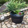 A wooden barrel planter with a water lily and a pink water lily flower in a garden setting with cobblestones and plants. Náhled obrázku produktu
