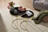 A set of nesting coffee tables with black glass tops and gold metal frames, styled with a vase of flowers, a candle, a teacup, and a blanket. Produktbild-Vorschau