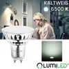 A woman is working at a desk in a room with a spotlight on the wall. A light bulb is in the foreground. Produktbild-Vorschau