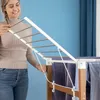 A woman is shown adjusting a white and silver folding drying rack with wooden accents. Several blue towels are hanging on the rack. Náhled obrázku produktu
