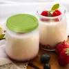 Two jars of yogurt with fruit on a wooden surface, with a white cloth and flowers in the background. Náhľad obrázku produktu