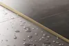 A close-up view of a gold-colored profile strip separating two dark gray tiled surfaces, with water droplets scattered on the tiles. Náhľad obrázku produktu