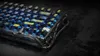 A close-up, angled view of a mechanical keyboard with transparent black casing and illuminated blue and yellow keys. Produktbild-Vorschau