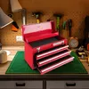 A red toolbox with three drawers open, sitting on a green cutting mat on a wooden workbench. Tools and other items are visible on the pegboard wall behind the toolbox. Náhled obrázku produktu