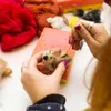 A person's hands are shown needle felting a small animal figure. Various colors of wool roving and sponges are visible in the background. Produktbild-Vorschau