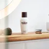 A bottle of Tabac Original aftershave lotion is displayed on a wooden shelf, alongside a shaving brush and a safety razor. Produktbild-Vorschau