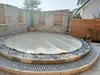 A round pool cover is shown in an outdoor setting, surrounded by stone walls and wooden fencing. Náhled obrázku produktu