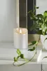 A silver LED candle with a flickering flame sits on a white marble surface next to a green plant. Podgląd zdjęcia produktu