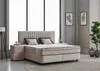 A bedroom scene featuring a beige boxspring bed with a tall, fluted headboard, two nightstands, and decorative items. Produktbild-Vorschau