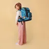A young person wearing a beanie and pink pants stands with a large blue backpack on their back, looking over their shoulder at the camera. Produktbild-Vorschau