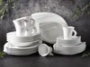 A collection of white porcelain dinnerware, including plates, bowls, and cups, arranged on a dark surface with a textured gray background and some greenery. Produktbild-Vorschau