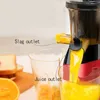 A close-up view of a juicer in operation, with juice flowing into a glass and pulp exiting another outlet. Oranges and bread are visible in the background. Náhled obrázku produktu