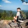A young man sits on a wooden bench overlooking a scenic mountain landscape, wearing a green backpack and holding hiking poles. Náhled obrázku produktu