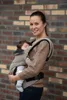 A woman is holding a baby in a carrier in front of a brick wall. Náhľad obrázku produktu
