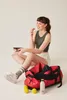 A woman is sitting on the floor, eating from a lunchbox, with a gym bag and sports equipment around her. Produktbild-Vorschau