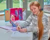 A young girl is sitting at a table, working on a craft project with a Barbie fashion salon and doll. Produktbild-Vorschau