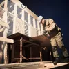 A construction worker is cutting a piece of wood on a table in front of a wooden structure. Produktbild-Vorschau