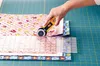 A hand is using a rotary cutter to cut fabric on a cutting mat. Produktbild-Vorschau