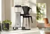 A coffee maker sits on a countertop next to a potted plant, with a blurred background of a living room. Produktbild-Vorschau