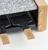 A close-up view of a raclette grill, showing the granite stone top and two of the non-stick pans with wooden handles. Produktbild-Vorschau