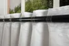 A close-up view of white curtains hanging on a black curtain rod in front of a window with greenery visible outside. Produktbild-Vorschau