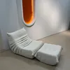 A modern beige lounge chair and ottoman are placed against a white wall with an orange accent light fixture. Produktbild-Vorschau