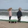 Two children stand on a beach, looking out at the ocean, with a green wagon in front of them. Produktbild-Vorschau