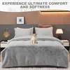 A cat sits on a plush gray blanket on a bed, with pillows and a bedside table visible. Produktbild-Vorschau