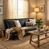 A living room scene featuring a black sofa with decorative pillows and a throw blanket, a wooden coffee table with candles and a plant, and a floor lamp. Produktbild-Vorschau