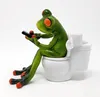 A decorative frog figurine sitting on a toilet, holding a remote control. Produktbild-Vorschau