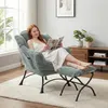 A woman is sitting in a comfortable armchair with a footrest, reading a magazine. The room has a large window, a wooden cabinet with plants and books, and a rug. Náhled obrázku produktu