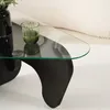 A close-up of a modern coffee table with a glass top and a black, sculptural base. A green glass is placed on the table. Produktbild-Vorschau