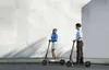 Two people on electric scooters in front of a textured wall. Produktbild-Vorschau