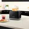 A black Russell Hobbs toaster with two slices of toast in it, sits on a white countertop next to a jar of jam and a plate with two slices of toast with jam. Podgląd zdjęcia produktu