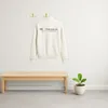 A cream-colored sweatshirt hangs on gold hooks above a wooden bench, with a potted plant to the right. Produktbild-Vorschau