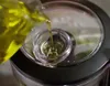 Oil is being poured from a glass container into a small glass bowl, which is placed on top of a kitchen appliance. Náhled obrázku produktu