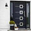 A black outdoor wall lamp is mounted on a white brick wall next to a dark blue front door. There are potted plants and a lantern on the doorstep. Náhled obrázku produktu