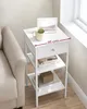 A white bedside table with a drawer and two shelves is shown next to a couch, near a window. The table has a mug, a framed picture, and books on it. Náhled obrázku produktu