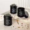 Three black storage canisters labeled 'Sugar', 'Coffee', and 'Tea' are arranged on a table with a beige cloth, alongside a small gold spoon and scattered dried flower petals. Produktbild-Vorschau