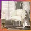 A cream-colored hanging chair with cushions is shown in a room with blinds and a side table. Náhled obrázku produktu