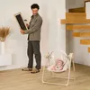 A man is holding a framed picture and a hammer, looking at a baby in a swing. A wooden staircase is in the background. Produktbild-Vorschau
