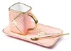 A pink ceramic coffee cup and saucer set with gold accents and two spoons, presented on a white background. Produktbild-Vorschau