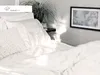 A close-up view of a neatly made bed with white linens and a decorative pillow, bathed in soft sunlight. A potted plant and a framed picture are visible on a shelf in the background. Produktbild-Vorschau