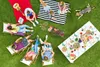 A group of people and a dog are enjoying a picnic on a grassy lawn, with a table laden with food and a grill nearby. Produktbild-Vorschau
