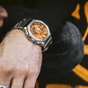 A close-up shot of a person's wrist wearing a silver and orange Casio G-Shock watch. Produktbild-Vorschau