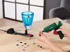 A person is using a green and red hot glue gun to attach decorative gems to a blue glass vase, with a charging station and scattered gems on a wooden table. Produktbild-Vorschau