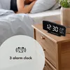 A digital alarm clock with temperature and humidity display sits on a wooden nightstand next to a bed where a person is sleeping. Produktbild-Vorschau
