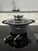 A black pot with a glass lid and two black pot holders are on a black countertop. The background shows a kitchen sink and cabinets. Náhľad obrázku produktu