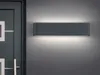 A modern, rectangular wall-mounted light fixture is shown mounted on a gray wall next to a dark gray door. Produktbild-Vorschau