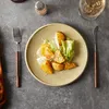A plate of food with roasted potato wedges and lettuce on a textured gray surface, with cutlery and a salt shaker. Produktbild-Vorschau